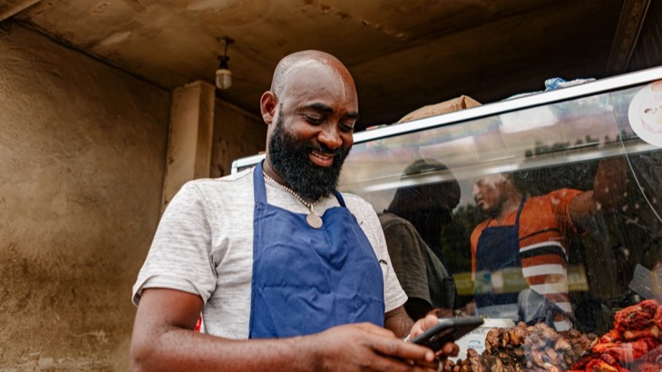 A local food vendor in an apron smiling at his phone, using a community chat app to message regular customers