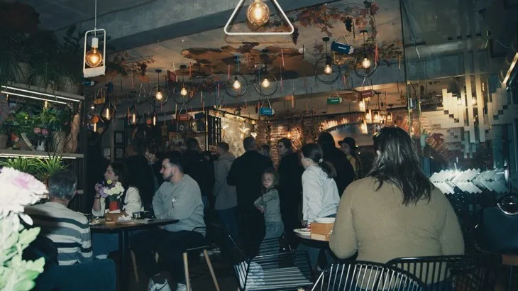 A busy neighborhood cafe full of people talking under warm hanging lights, an example of a local business operating as a third place during the friendship recession