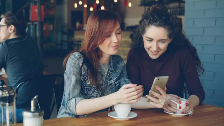 Two friends at a cafe looking at a phone together, checking in on a shared habit tracking streak