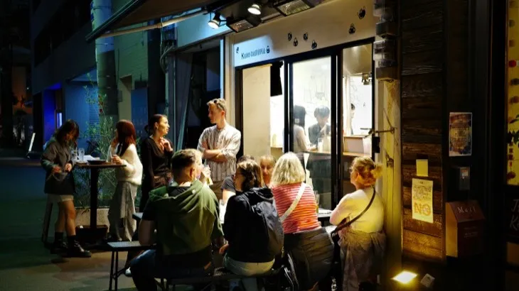 People gathered outside a lit restaurant at night, a recurring social habit in a neighborhood