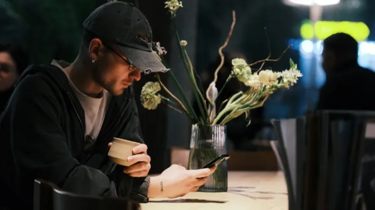 A man on his phone at a cafe table while other people sit nearby, an example of an autopilot social habit