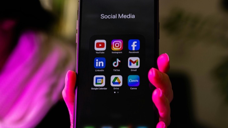 Hand holding a smartphone displaying social media app icons including YouTube, Instagram, Facebook, TikTok, and LinkedIn
